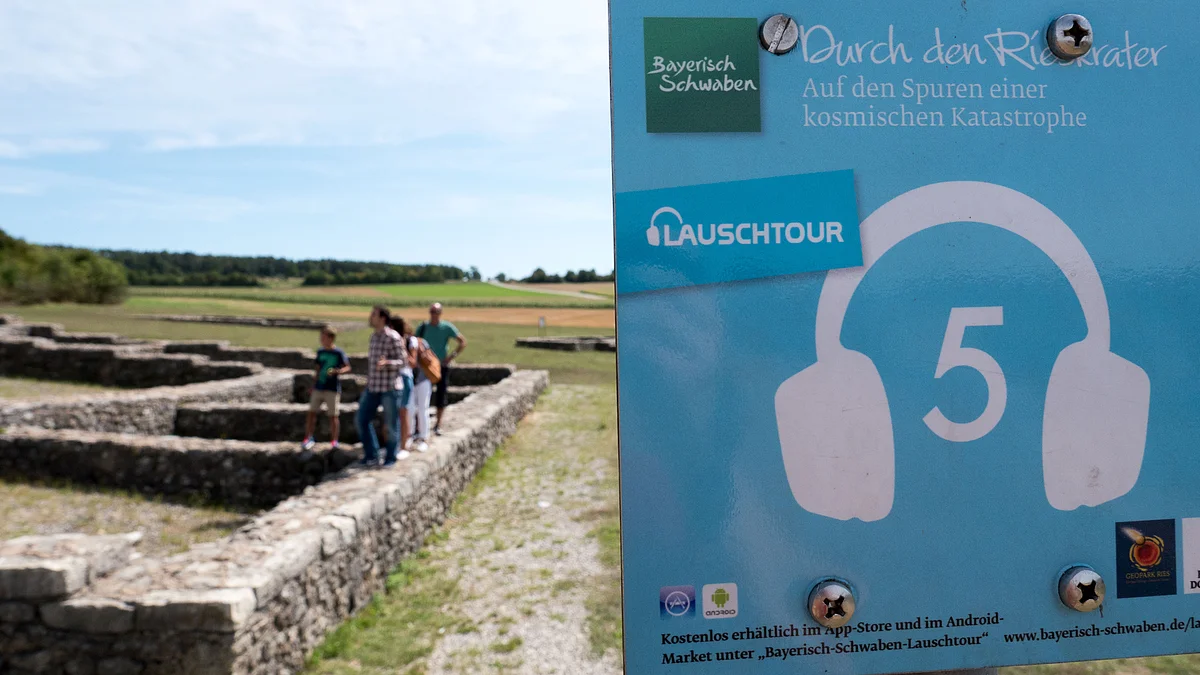 Lauschtour Rieskrater-Tour Ein kleines Schild weist auf die Lauschtour 5 "Durch den Rieskrater" hin. Im Hintergrund sind Personen zu sehen, die auf den Mauerfundamenten eines Gebäudes laufen.