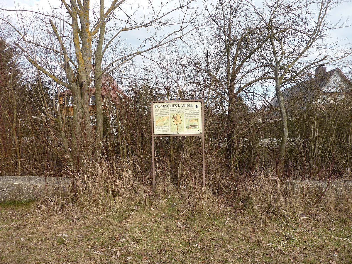 Vor einer Gruppe Büsche steht eine Informationstafel mit der Überschrift "Römisches Kastell".